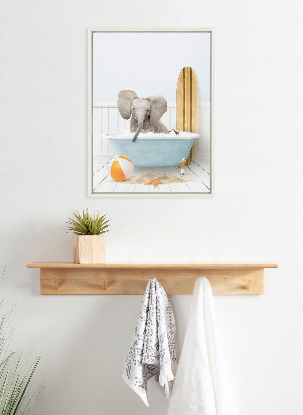 Framed canvas of a baby elephant sitting in a blue clawfoot bathtub with a wooden surfboard and a beach ball, displayed above a wooden wall shelf with hooks.