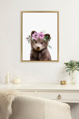 Framed canvas art of a young brown bear wearing a pink and purple flower crown, displayed on a white wall above a desk with decor and a plant in a glass vase.