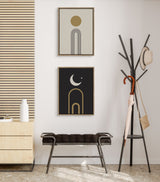 Two framed minimalist prints with geometric arches, one beige with gold sun above, one black with gold crescent moon and star, displayed above a black bench and next to a coat rack.