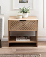 Compact wooden side table with chevron-patterned drawer front, black handle, open bottom shelf holding a magazine, and potted green plant on top.