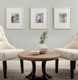 Three cream-colored Adlynn Beaded photo frames with black and white pictures hung on a soft green wall above a round wooden coffee table between two cream upholstered chairs.