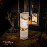 Tall cylindrical onyx table lamp with natural beige, brown, and white banding, placed on a textured wood surface reflecting in a nearby rectangular mirror.