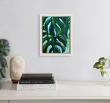 Framed 8x10 canvas featuring a close-up painting of dark green and turquoise monstera leaves against a soft blue background, hung on a light gray wall above a white surface.