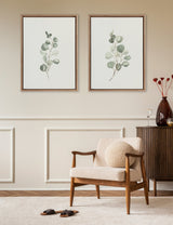 Two framed eucalyptus botanical prints with delicate green leaves on a beige wall above a mid-century modern wooden armchair with cream cushions and a round pillow.