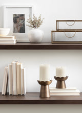 Two bronze metal pillar candle holders with scalloped edges holding white candles, placed on a dark wooden shelf with stacked white books beside and above.