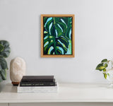 Wood-framed canvas featuring a close-up painting of large green monstera leaves with dark and light green highlights, hanging centered above a white surface with books and plants.
