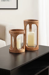 Two cylindrical wooden candle holders with curved arch cutouts, each containing a white pillar candle inside clear glass, placed on a black side table near a beige sofa.