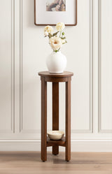 Tall, narrow round wood side table with dark brown finish, supporting a white vase with cream flowers, and a small bowl on the lower shelf against a paneled wall.
