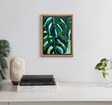 Small framed painting of large green tropical leaves with dark shadows and white highlights, hanging on a light gray wall above a white surface with books and plants.