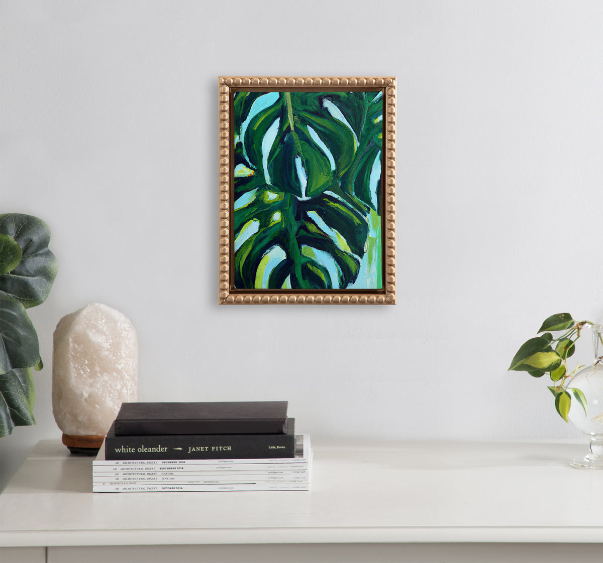 Small framed painting of large green tropical leaves with dark shadows and white highlights, hanging on a light gray wall above a white surface with books and plants.