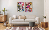 Two abstract canvas paintings with dynamic brushstrokes in pink, yellow, black, and blue tones, framed and hung side by side above a beige mid-century sofa.