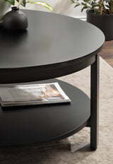 Close-up of a round black wood coffee table with a matte top and lower shelf holding a magazine, positioned on a light patterned rug with potted plants nearby.