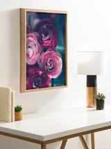 Framed floral art print of pink and dark magenta ranunculus flowers with green blurred background, hanging on white wall above a white desk with wood and black lamp.