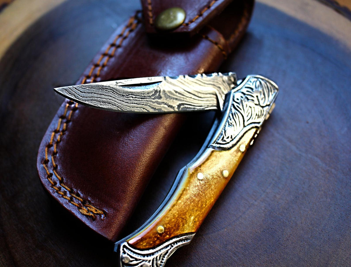 Damascus steel pocket knife with burnt bone handles and steel bolsters resting on a wooden surface