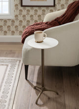 Round white marble side table with a tripod bronze metal base holding a beige coffee cup beside a cream upholstered armchair with a rust orange knit blanket.