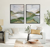 Two framed abstract canvas paintings featuring soft green and gray hills with misty white and beige skies, hung side by side above a white sofa in a modern living room.