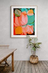 Abstract botanical art print featuring overlapping translucent leaf shapes in orange, teal, and green tones framed and hung on a textured light gray wall above a wooden table and potted plant.