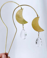 Gold-toned metal wire suncatcher with two crescent moon shapes, each dangling a faceted clear crystal prism, held against a white background.