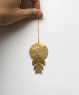 Hand holding a hanging ornament with four linked hammered gold metal shapes decreasing in size, resembling moon phases, against a plain white wall.