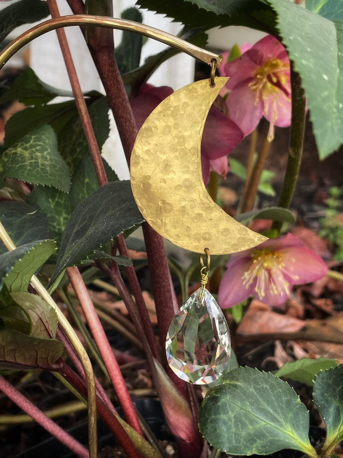 Hammered brass crescent moon attached to a curved brass rod with a hanging clear faceted crystal teardrop, set among green leaves and pink flowers outdoors.
