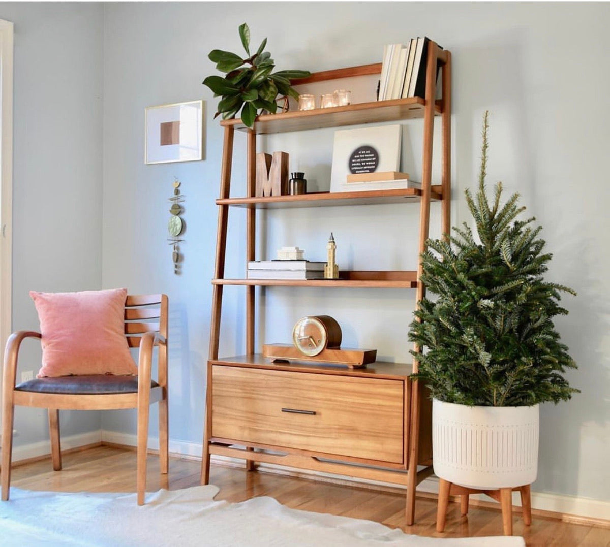 Wooden shelving unit with three open shelves and a closed cabinet base, styled with books, plants, and decor, next to a wooden chair with a pink cushion and a potted fir tree