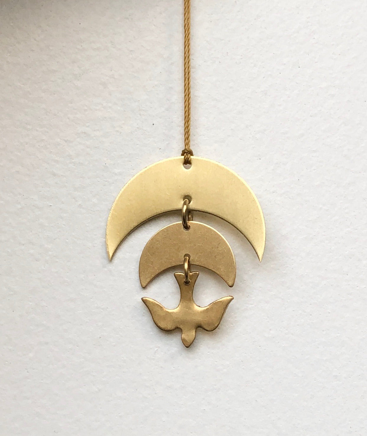 Brass ornament with three connected shapes: a large crescent at top, a smaller crescent in the middle, and a stylized bird figure hanging at the bottom on a braided cord.