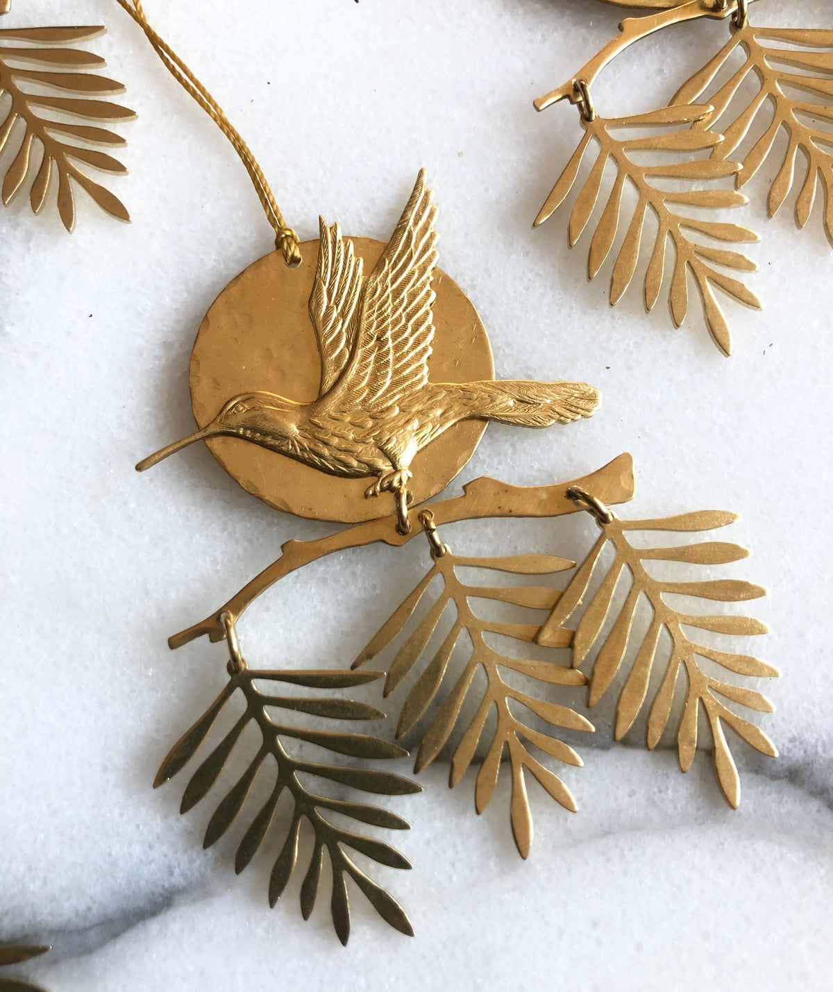 Brass hanging ornament featuring a detailed bird in flight in front of a circular disk, with three delicate palm fronds dangling below on a light background.