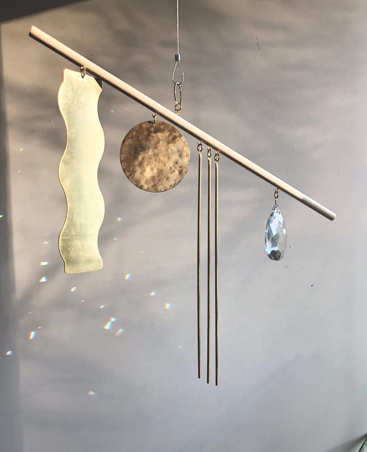 Horizontal wooden stick suspending a large wavy yellow translucent shape, a brown round metal disc, three thin gold chimes, and a teardrop-shaped clear crystal prism against a neutral background.