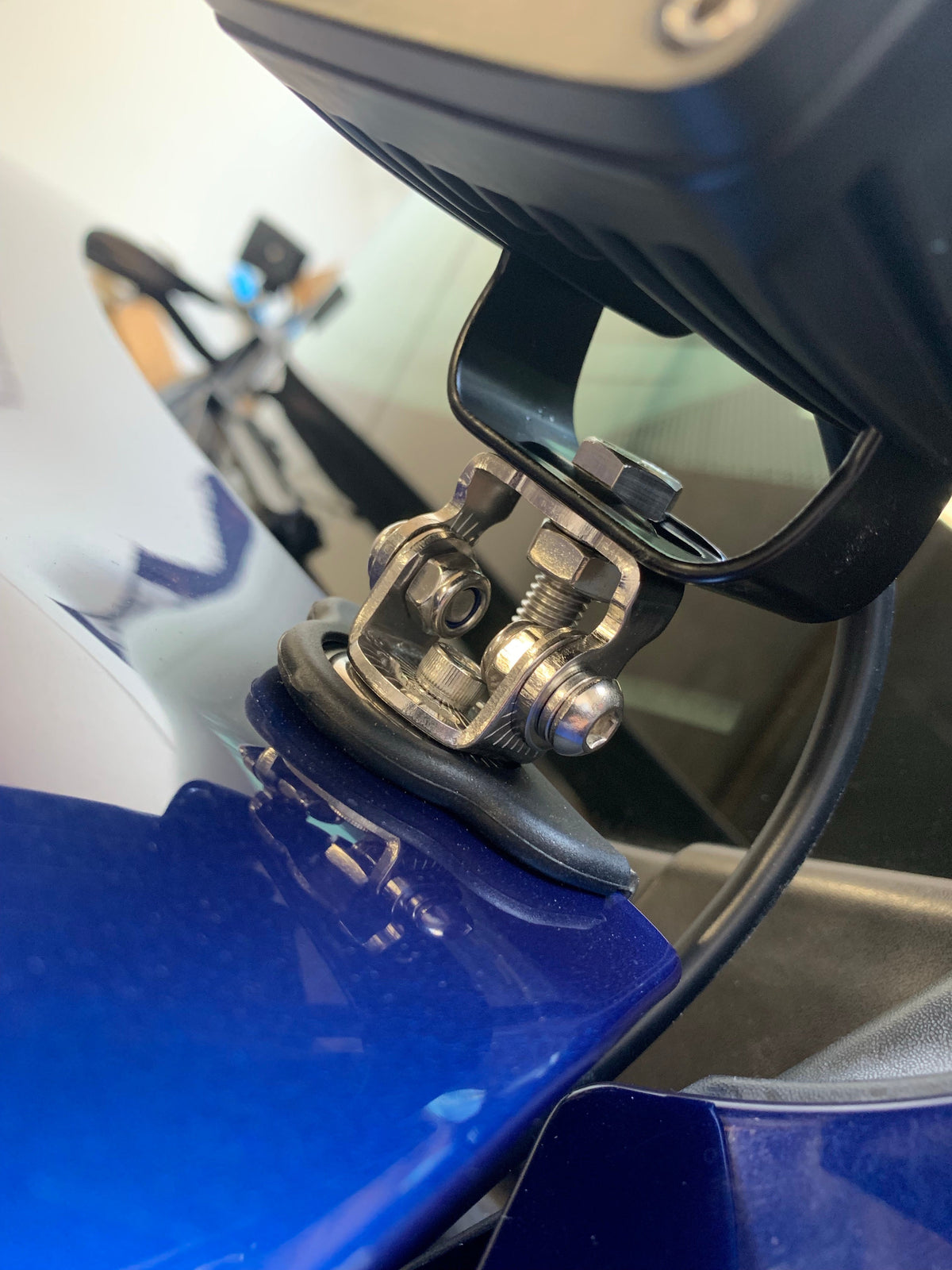 Close-up of a silver metal clamp securing a black ditch light to a blue vehicle surface, with a rubber gasket between clamp and surface and a black cable extending from the light.