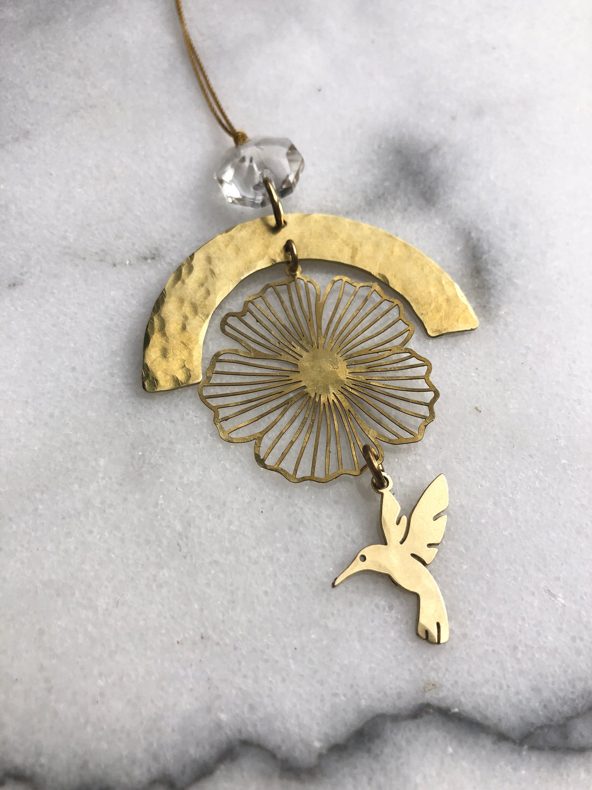 Gold-toned ornament featuring a hammered crescent arch, delicate wireframe flower, and a small hummingbird charm suspended below, with a vintage clear glass crystal bead at the top.
