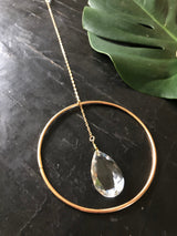 Clear teardrop crystal hanging from a thin gold chain inside a simple gold circular frame on a dark textured surface with a green leaf partially visible.