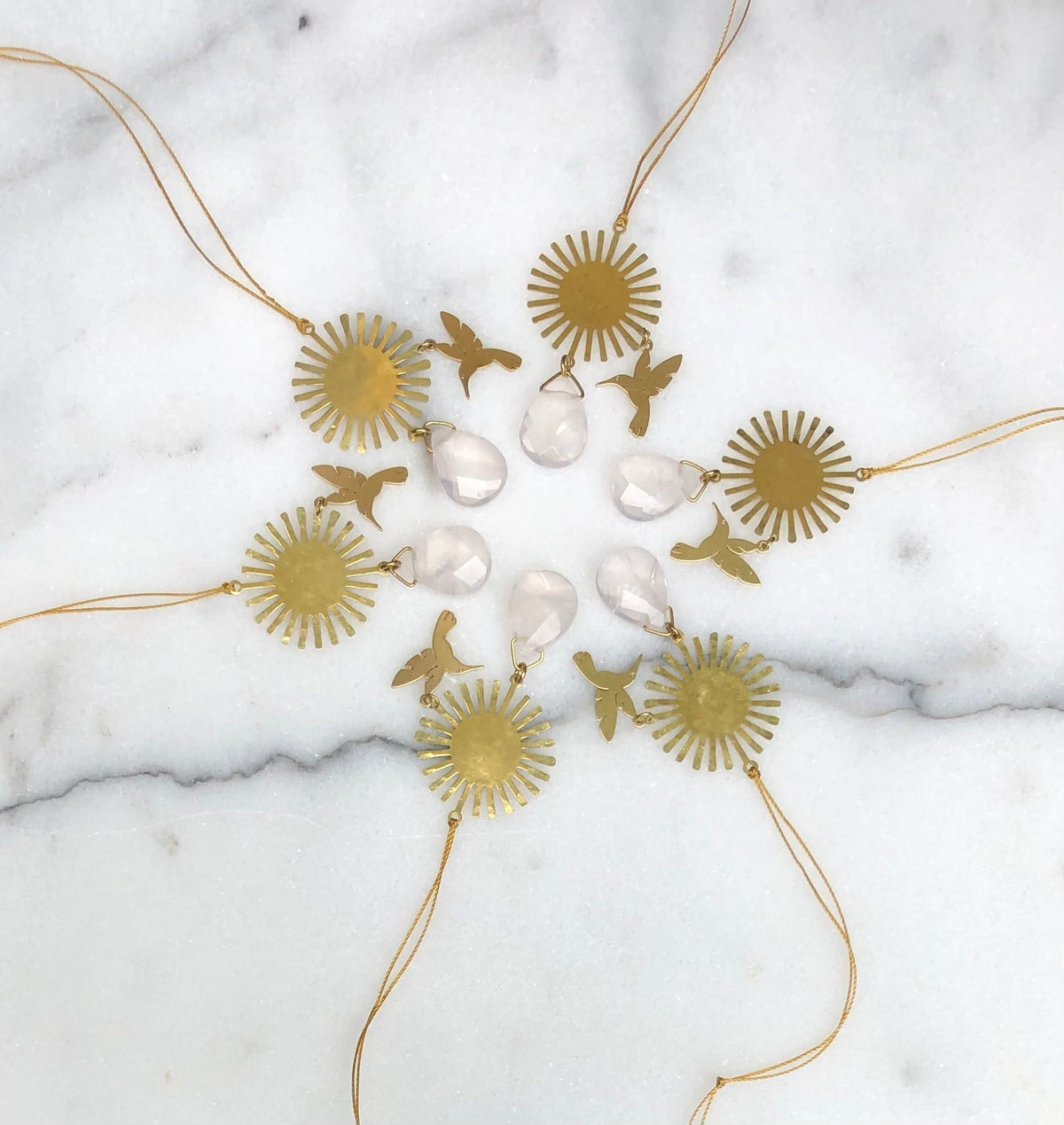 Circular gold sunburst ornaments paired with small gold hummingbird shapes and teardrop-shaped clear rose quartz stones, arranged in a circle on white marble.