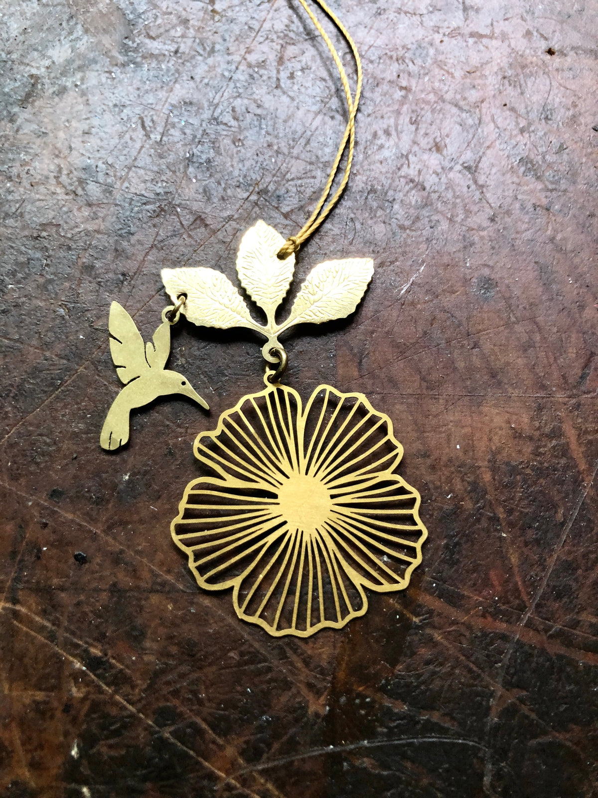 Delicate gold-colored ornament featuring a stylized hibiscus flower with radial petal lines and a hovering hummingbird near three textured leaves, resting on a worn brown surface.