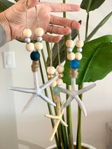 Three hanging starfish ornaments with natural wooden beads and blue accent beads, held in hand against a background of green plant leaves.