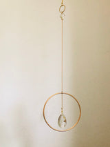 Delicate gold-tone circular frame hanging vertically with a clear, faceted teardrop crystal suspended inside against a plain light background.