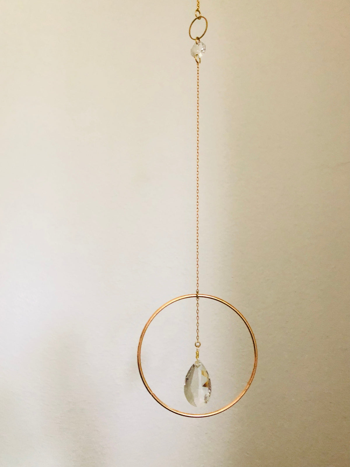Delicate gold-tone circular frame hanging vertically with a clear, faceted teardrop crystal suspended inside against a plain light background.