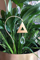 Gold triangular suncatcher with a clear teardrop crystal hanging inside, suspended on a curved gold metal rod among green leafy plants near a window.
