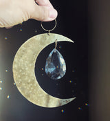 Hand holding a crescent moon-shaped brass suncatcher with a hanging faceted clear teardrop crystal, casting small light reflections on a dark background.