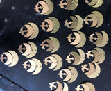 Multiple brass crescent moon ornaments with a small bird shape cutout, arranged on a dark textured surface with visible rivets.