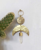Gold-toned metal pendant with hammered textures featuring a circle and crescent moon shapes, topped by a ring and dangling a clear quartz crystal point, beside a cactus.