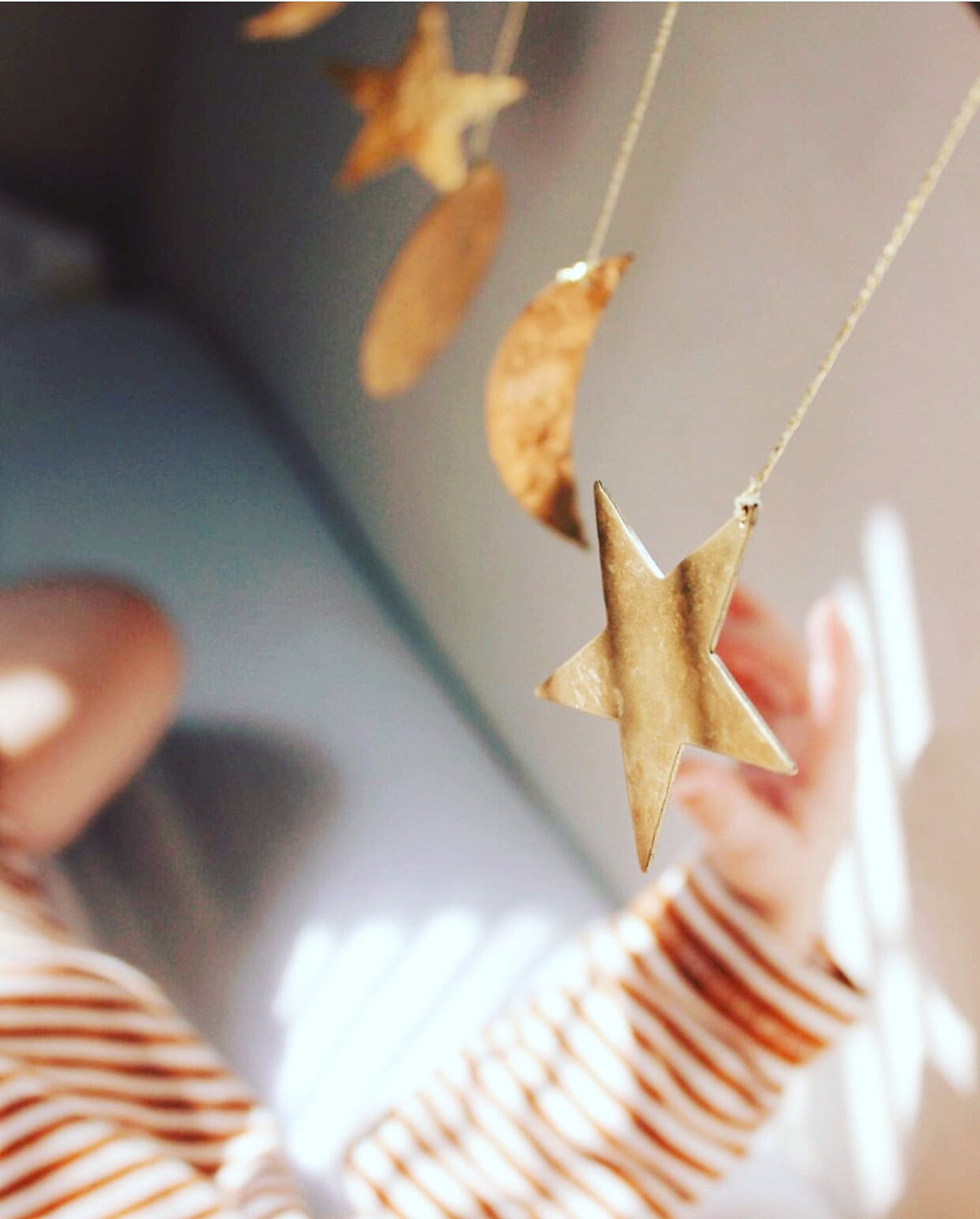 Close-up of gold hammered brass ornaments in star, crescent moon, and circular shapes hanging from strings against a softly lit background with a striped sleeve reaching towards them