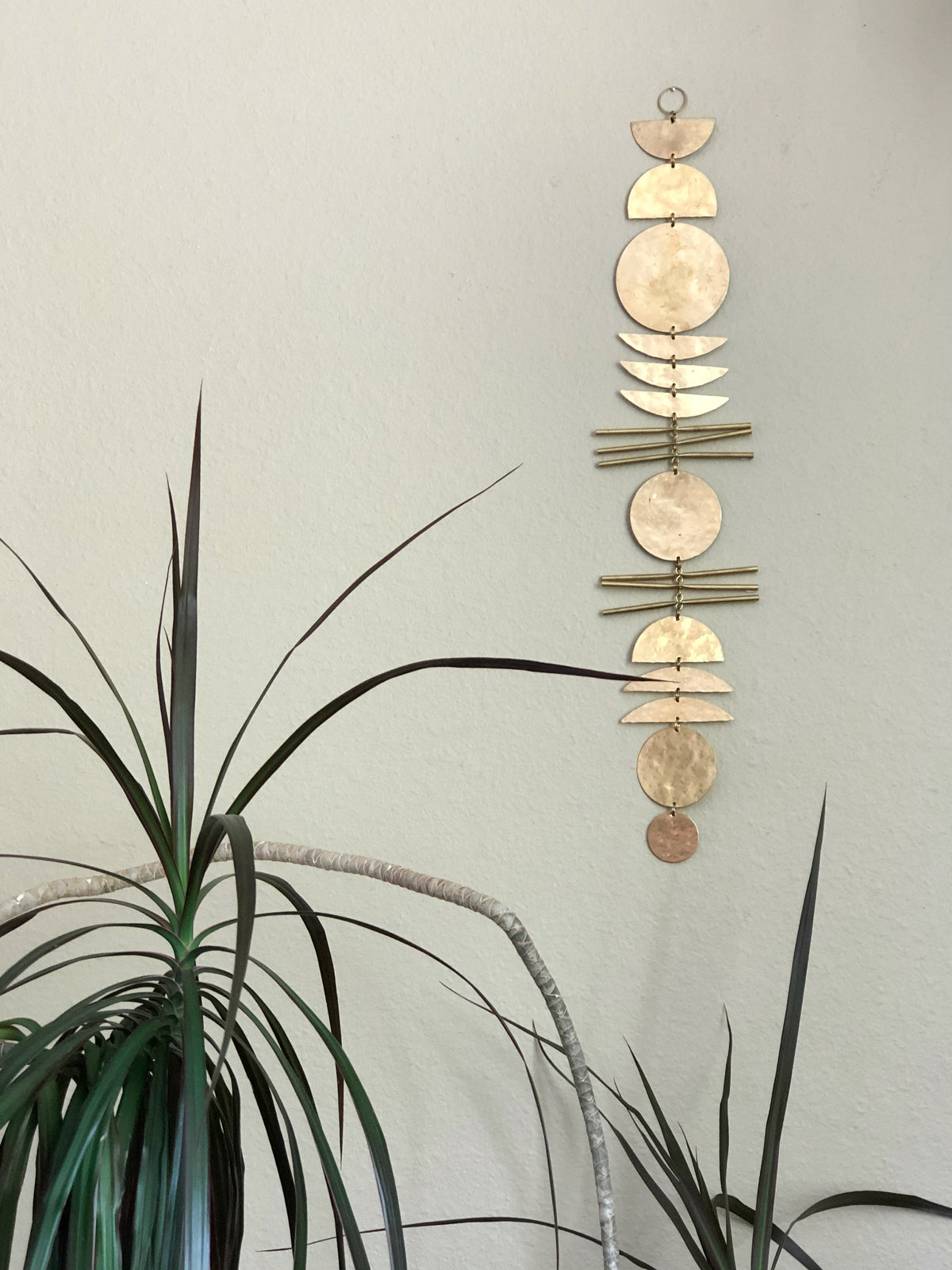 Vertical wall hanging with gold-toned geometric discs and rods arranged in a celestial-inspired pattern, displayed on a light gray wall beside tall, pointed green leaves.