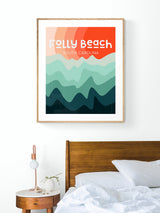 Modern art print above bed featuring stylized abstract waves in layers of teal and green below a gradient orange sky with text 'Folly Beach South Carolina' in white.