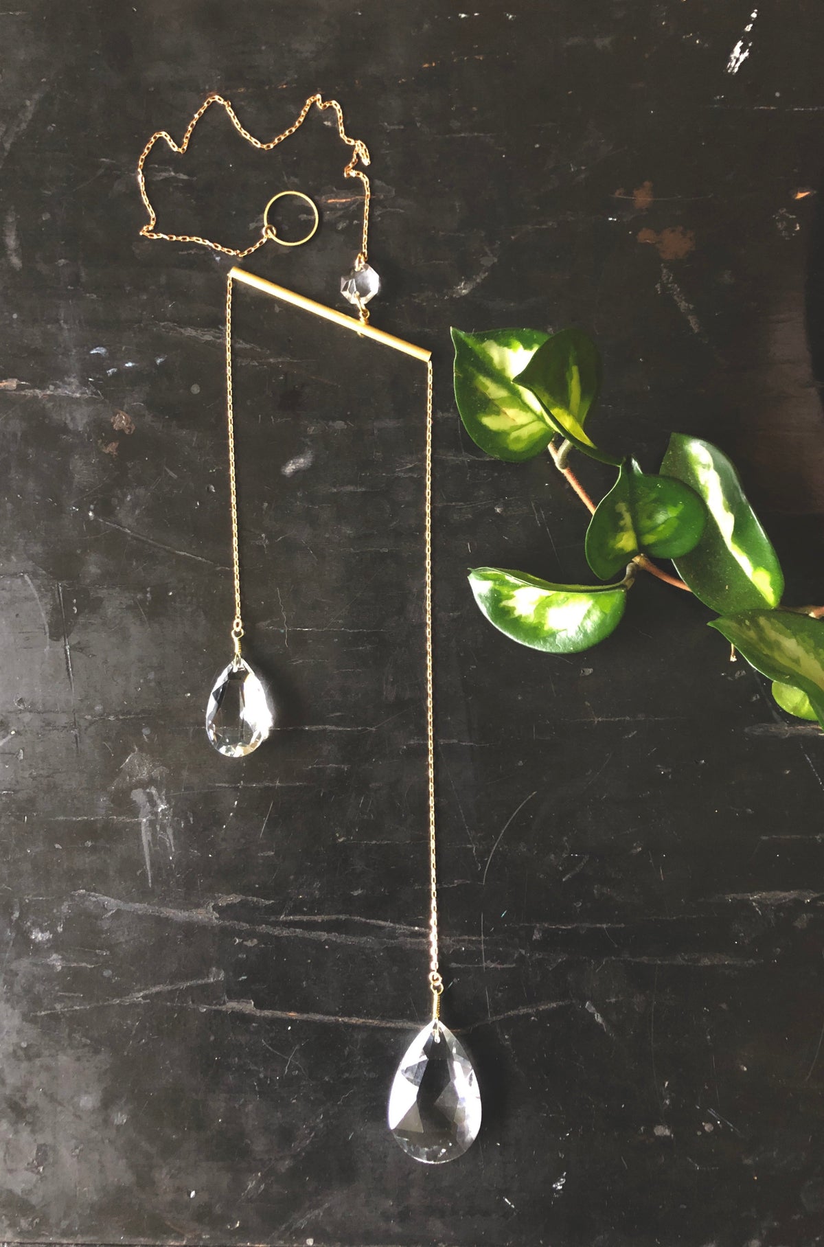 Delicate metal wire suncatcher shaped like a chicken with two faceted glass teardrop crystals hanging from thin chains, displayed on a dark surface beside a green leafy plant.