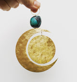 Hand holding a decorative piece with a hammered gold crescent moon and a suspended hammered gold circle below a turquoise stone bead attached to a small gold ring.