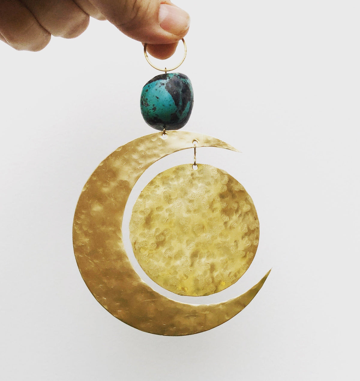 Hand holding a decorative piece with a hammered gold crescent moon and a suspended hammered gold circle below a turquoise stone bead attached to a small gold ring.