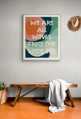 Abstract art print with layered, rounded shapes in green, orange, and cream colors and text reading 'We are all waves from the same sea' hung above a wooden bench with a white cloth draped over it.