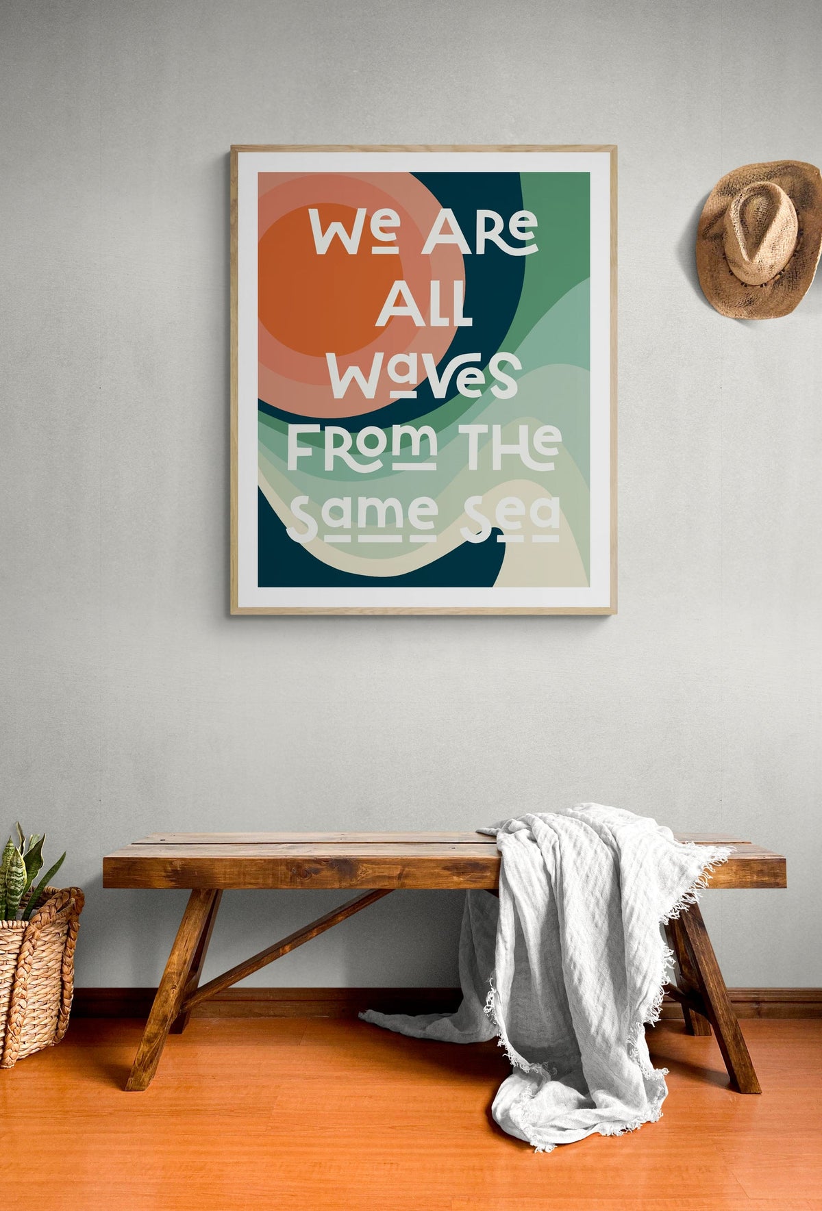 Abstract art print with layered, rounded shapes in green, orange, and cream colors and text reading 'We are all waves from the same sea' hung above a wooden bench with a white cloth draped over it.