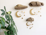 Three driftwood pieces on a white wall, each with hanging gold crescent moons and stars in varying sizes, beside a green houseplant with variegated leaves.