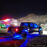 Black pickup truck with blue underglow lights parked on rocky terrain at night near other vehicles and an American flag, with hills in the background.